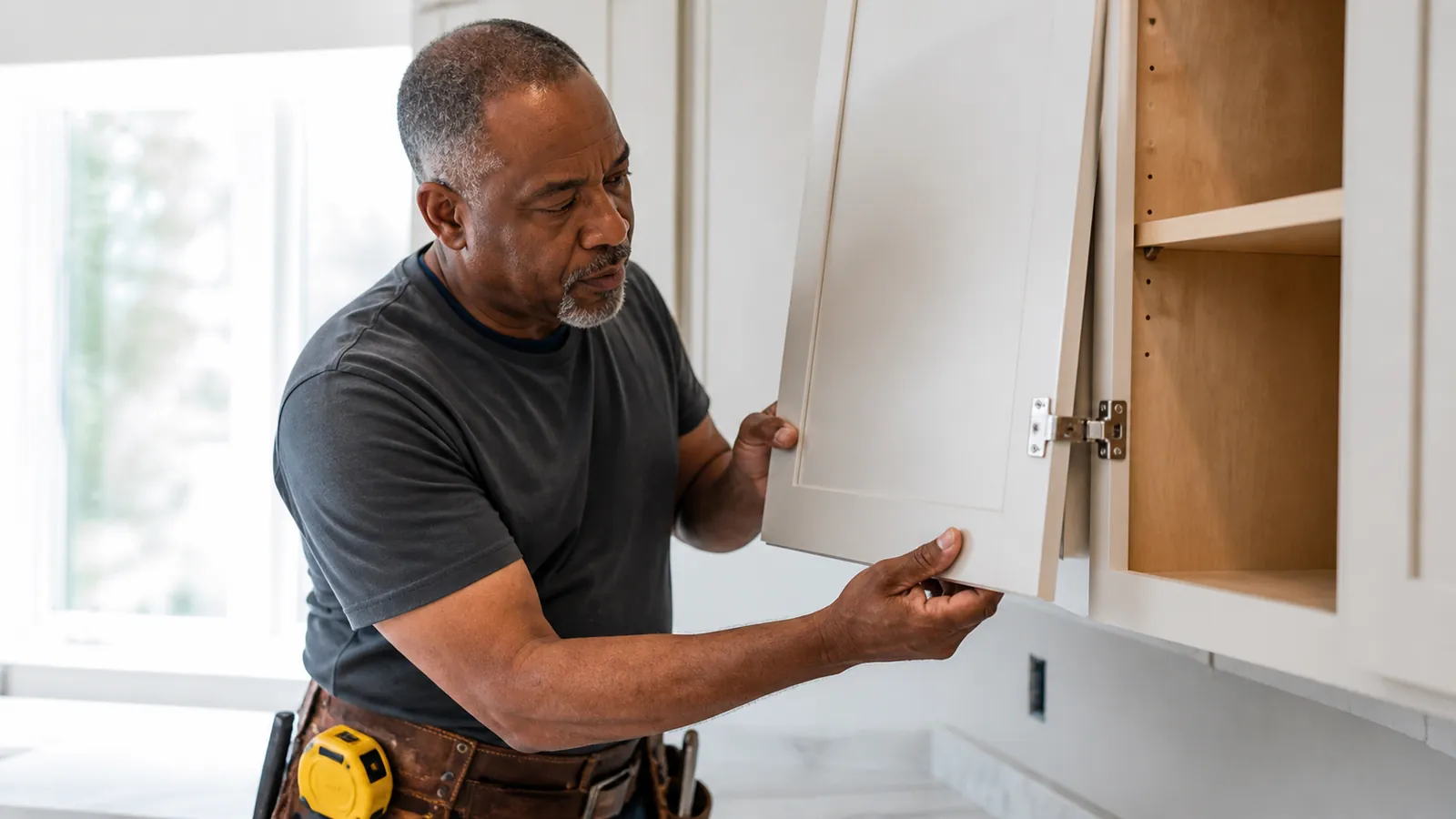Handyman fitting a new cabinet door in a bright kitchen, yellow tape measure on his tool belt