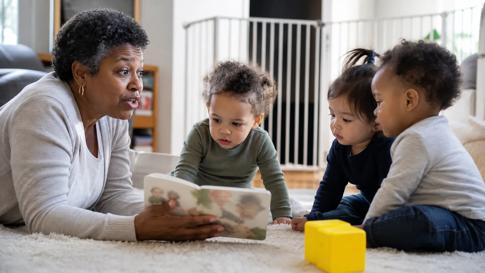 Home daycare provider reading to three toddlers on a rug in a bright gated living room
