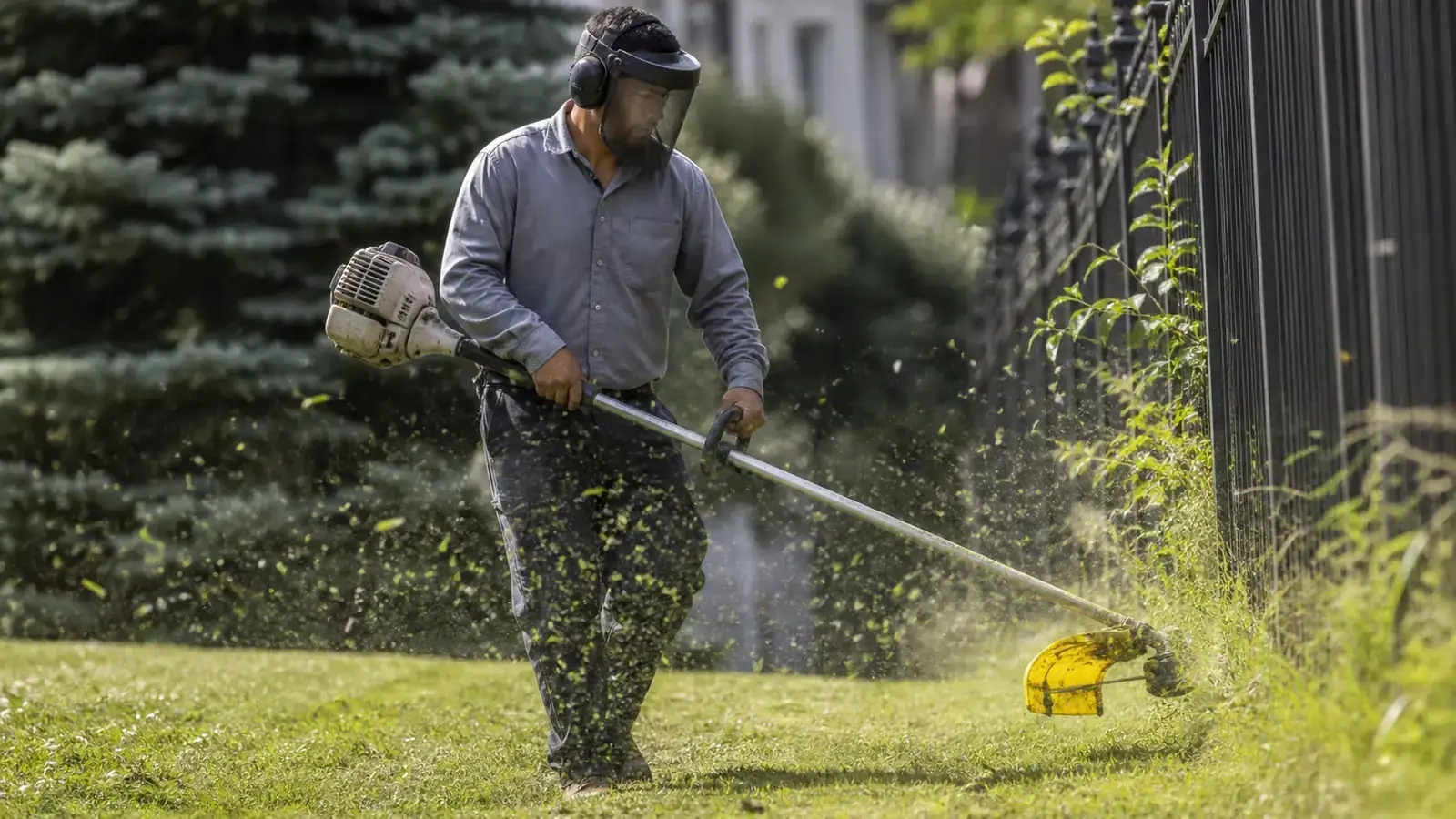 Crew member in full PPE trimming a fence line, yellow trimmer guard catching the light